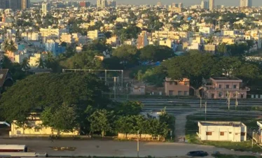 An aerial view of a red passenger train parked at a railway station in Bangalore, with a sprawling cityscape and distant high-rise buildings under a dramatic sky.
