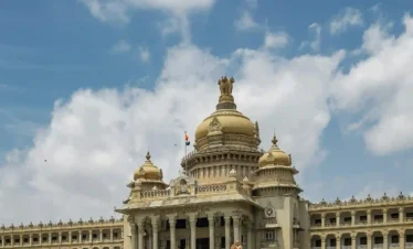 The Vidhana Soudha in Bengaluru, a massive Neo-Dravidian style stone building with a central gold-colored dome under a bright blue sky with white clouds.