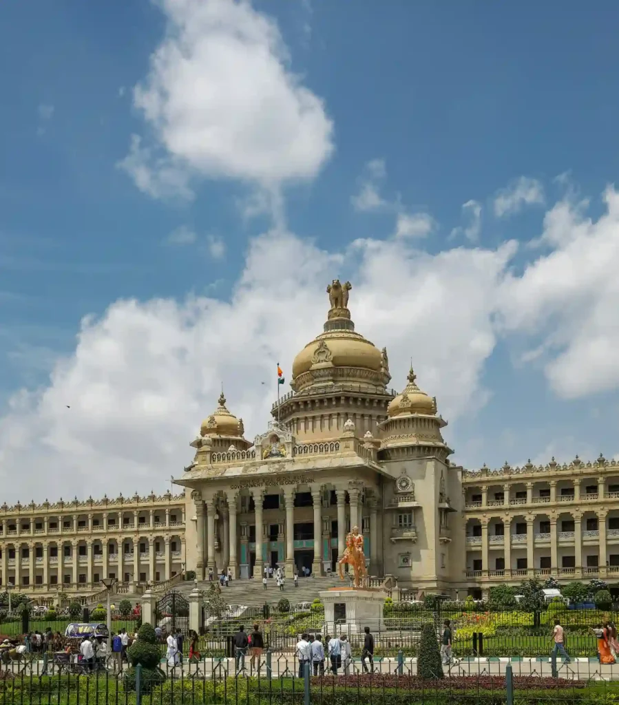 The Vidhana Soudha in Bengaluru, a massive Neo-Dravidian style stone building with a central gold-colored dome under a bright blue sky with white clouds.