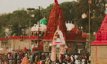A vertical, high-angle shot of a crowded riverside ghat in India, featuring traditional Hindu temples with distinct red and white spires (shikharas). Numerous devotees are gathered on the stone steps leading into the water, some bathing and others standing in prayer. The scene is bathed in a warm, golden sunset glow, highlighting the ancient architecture and the spiritual energy of the location.