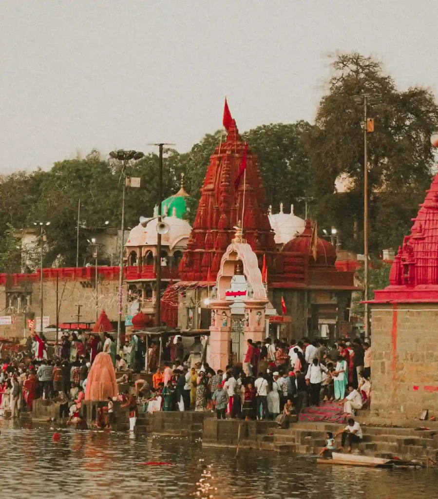 A vertical, high-angle shot of a crowded riverside ghat in India, featuring traditional Hindu temples with distinct red and white spires (shikharas). Numerous devotees are gathered on the stone steps leading into the water, some bathing and others standing in prayer. The scene is bathed in a warm, golden sunset glow, highlighting the ancient architecture and the spiritual energy of the location.
