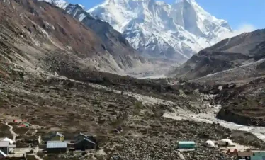 A wide, high-angle shot of a high-altitude Himalayan valley. In the foreground, a small mountain village with stone and metal-roofed buildings is scattered across a rocky, brown landscape. In the background, massive, snow-covered mountain peaks rise sharply against a clear, deep blue sky. A narrow river or glacial path winds through the valley floor toward the base of the mountains.