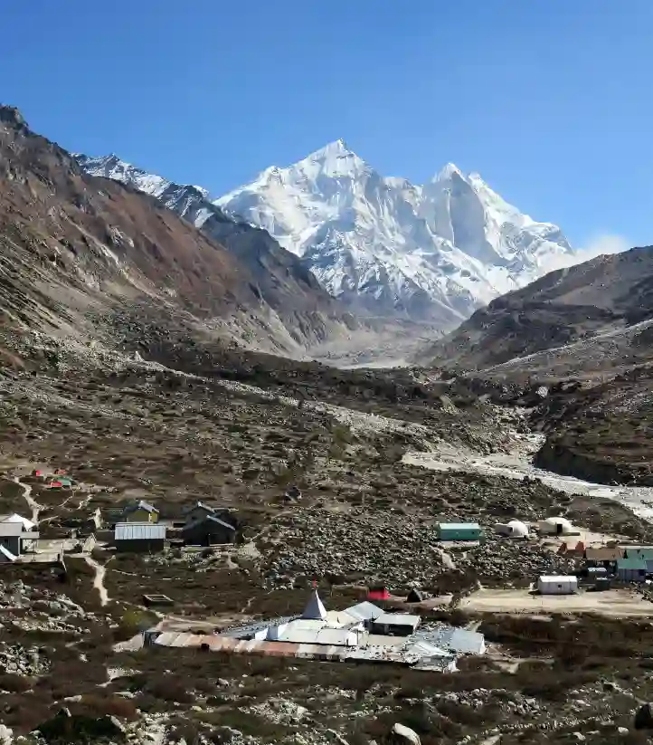 A wide, high-angle shot of a high-altitude Himalayan valley. In the foreground, a small mountain village with stone and metal-roofed buildings is scattered across a rocky, brown landscape. In the background, massive, snow-covered mountain peaks rise sharply against a clear, deep blue sky. A narrow river or glacial path winds through the valley floor toward the base of the mountains.