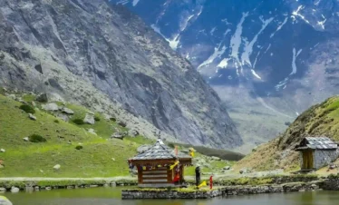 A scenic view of the Yulla Kanda trek featuring a traditional wooden Hindu temple built on a small stone island in the center of a calm alpine lake. Lush green meadows line the foreground, while a dramatic backdrop of towering, rugged gray mountains with patches of snow rises into a blue sky.