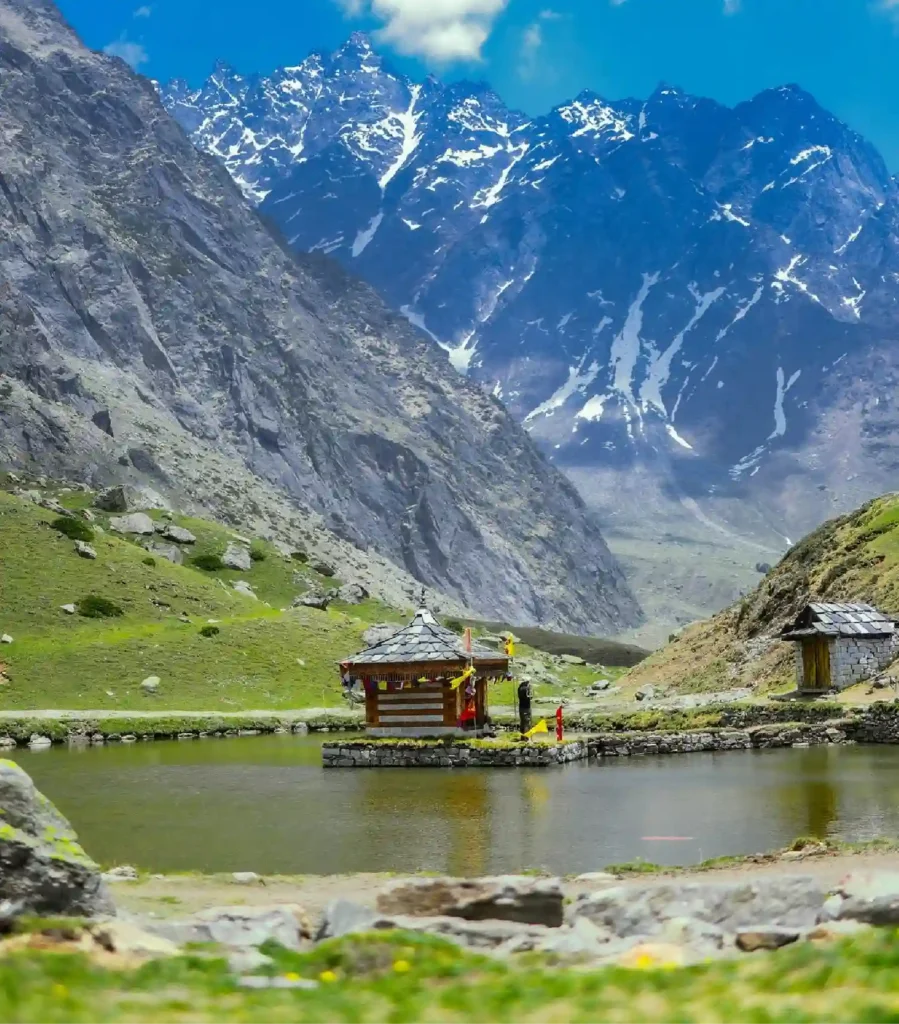 A scenic view of the Yulla Kanda trek featuring a traditional wooden Hindu temple built on a small stone island in the center of a calm alpine lake. Lush green meadows line the foreground, while a dramatic backdrop of towering, rugged gray mountains with patches of snow rises into a blue sky.