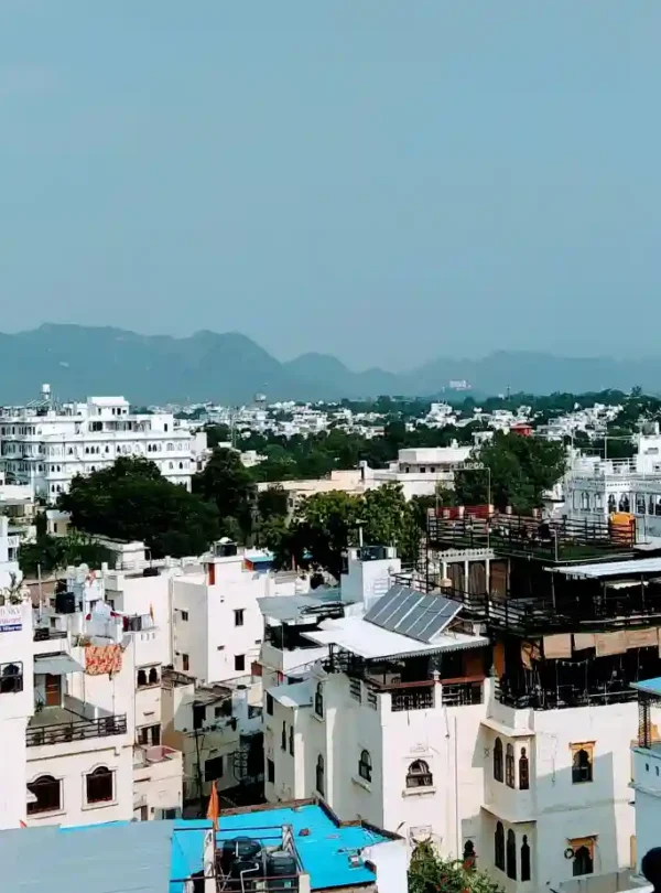 Panoramic view of whitewashed buildings and rooftops in Udaipur with Aravalli hills in the background under a clear sky.