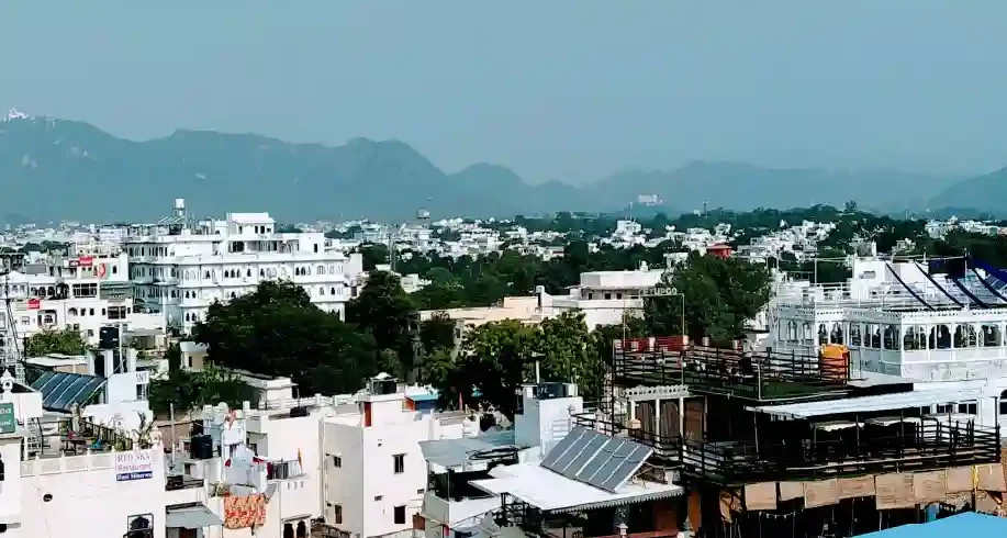 Panoramic view of whitewashed buildings and rooftops in Udaipur with Aravalli hills in the background under a clear sky.
