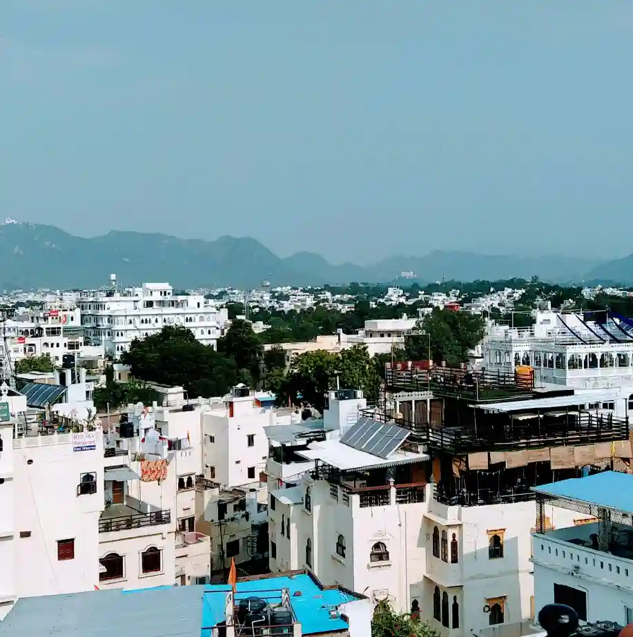 Panoramic view of whitewashed buildings and rooftops in Udaipur with Aravalli hills in the background under a clear sky.