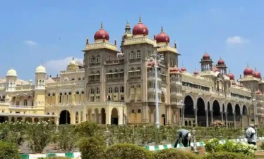 A bright, daytime view of the Mysore Palace with its ornate facade and red domes, seen from across the surrounding green gardens.