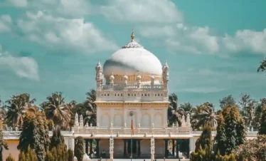 A frontal view of the Gumbaz near Mysore, featuring a large central white dome and ornate carvings. A stone walkway lined with manicured trees leads groups of visitors toward the entrance under a cloudy turquoise sky.
