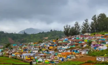 A wide shot of a multi-colored village nestled on a lush green hillside under a moody, overcast sky. The houses are painted in a vibrant patchwork of blues, yellows, pinks, and reds. In the foreground, neat terraced farm plots transition into the residential area, while tall eucalyptus trees and mist-covered mountains define the horizon.
