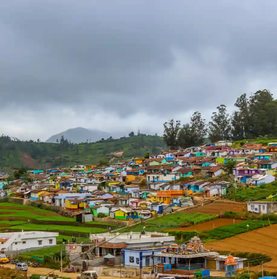 A wide shot of a multi-colored village nestled on a lush green hillside under a moody, overcast sky. The houses are painted in a vibrant patchwork of blues, yellows, pinks, and reds. In the foreground, neat terraced farm plots transition into the residential area, while tall eucalyptus trees and mist-covered mountains define the horizon.