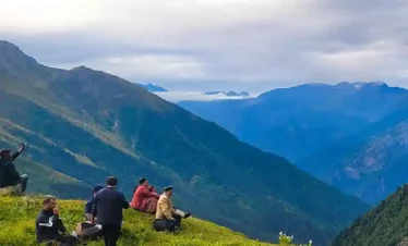 A group of trekkers resting on a grassy green hillside overlooking vast mountain valleys on the Yulla Kanda trek under an overcast sky.