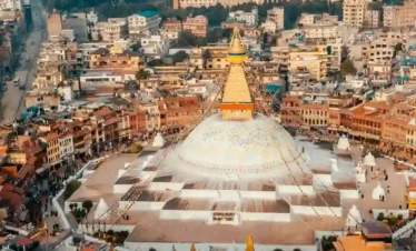 An aerial shot of the iconic Boudhanath Stupa in Kathmandu, showing its grand white dome and golden spire centered within a bustling circular market and residential area.