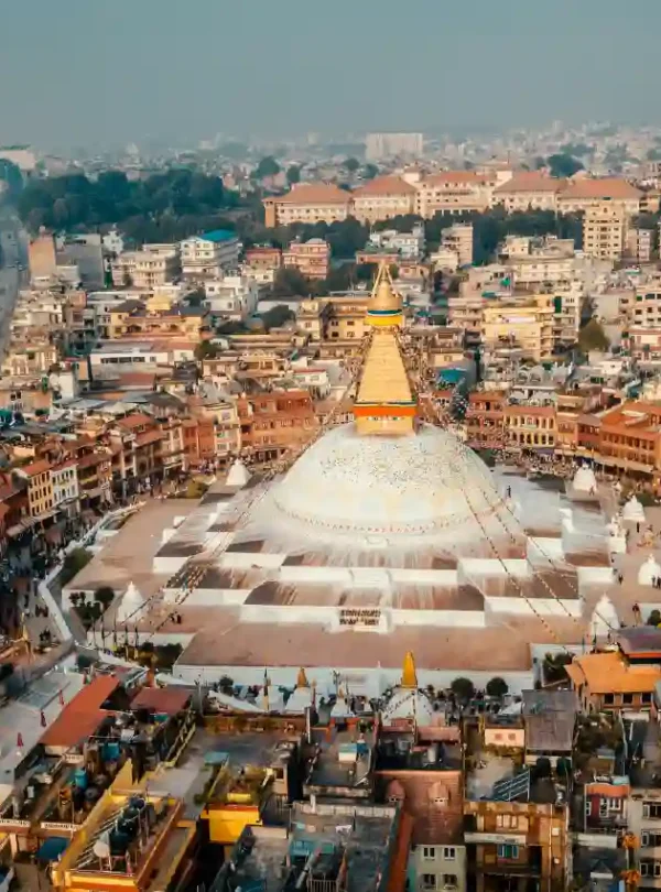 An aerial shot of the iconic Boudhanath Stupa in Kathmandu, showing its grand white dome and golden spire centered within a bustling circular market and residential area.