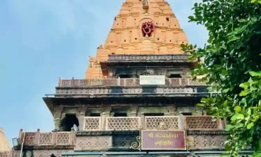 A low-angle shot of the Mahakaleshwar Temple's main spire (shikhara) in Ujjain. The lower section features dark, intricately carved stone pillars and balconies with a sign reading "Shree Mahakaleshwar Jyotirlinga" in Hindi. Above, a large, orange-toned, tiered pyramid spire rises toward a blue sky, adorned with a red "Om" symbol and a fluttering red flag at the very peak. Green tree branches frame the right side of the image.