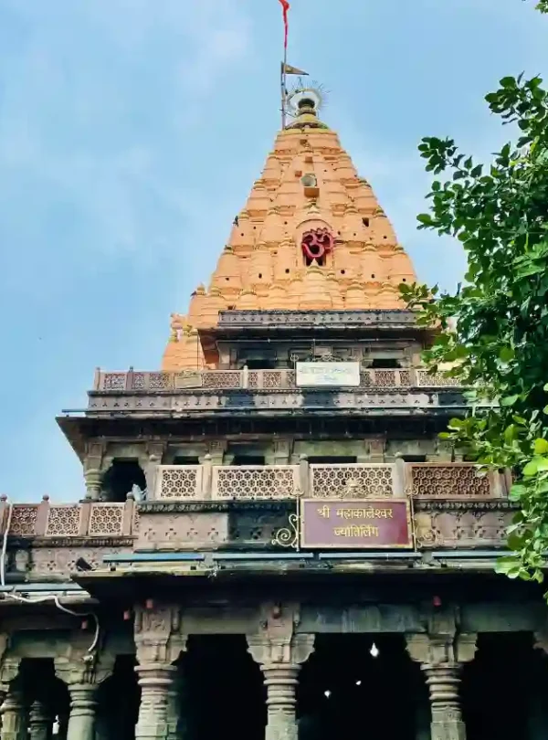 A low-angle shot of the Mahakaleshwar Temple's main spire (shikhara) in Ujjain. The lower section features dark, intricately carved stone pillars and balconies with a sign reading "Shree Mahakaleshwar Jyotirlinga" in Hindi. Above, a large, orange-toned, tiered pyramid spire rises toward a blue sky, adorned with a red "Om" symbol and a fluttering red flag at the very peak. Green tree branches frame the right side of the image.