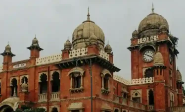 A majestic Indo-Saracenic style heritage building made of red sandstone and white marble. The structure features grand domes, intricate scalloped arches, and a prominent four-faced clock tower on the right. Lush green trees and a paved courtyard are visible in the foreground under an overcast, grey sky.