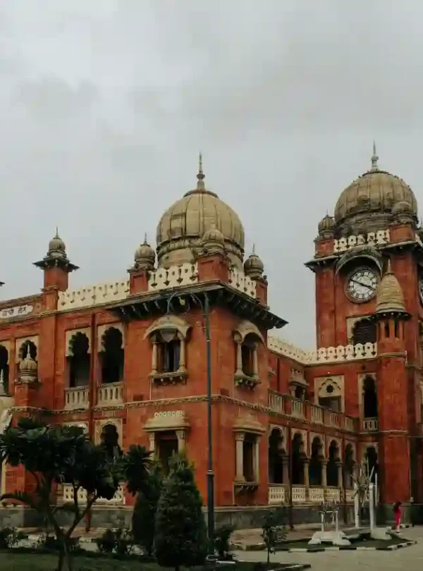 A majestic Indo-Saracenic style heritage building made of red sandstone and white marble. The structure features grand domes, intricate scalloped arches, and a prominent four-faced clock tower on the right. Lush green trees and a paved courtyard are visible in the foreground under an overcast, grey sky.
