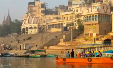 A vibrant orange tourist boat on the Ganges River in Varanasi, with seagulls in the water and the historic stone ghats and temples of Kashi rising in the background.