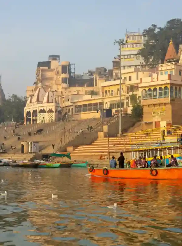 A vibrant orange tourist boat on the Ganges River in Varanasi, with seagulls in the water and the historic stone ghats and temples of Kashi rising in the background.
