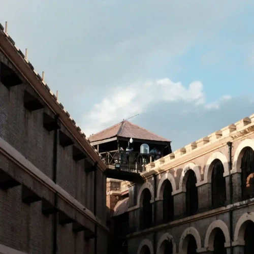 A low-angle shot of the Cellular Jail (Kala Pani) in the Andaman Islands, highlighting its distinct colonial architecture. The image shows the multi-tiered brick corridors with large arches on the right, a plain prison wing on the left, and the central wooden watchtower bridging them under a bright sky.