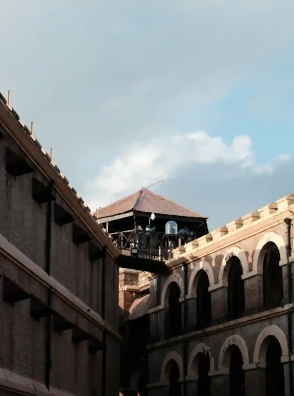 A low-angle shot of the Cellular Jail (Kala Pani) in the Andaman Islands, highlighting its distinct colonial architecture. The image shows the multi-tiered brick corridors with large arches on the right, a plain prison wing on the left, and the central wooden watchtower bridging them under a bright sky.