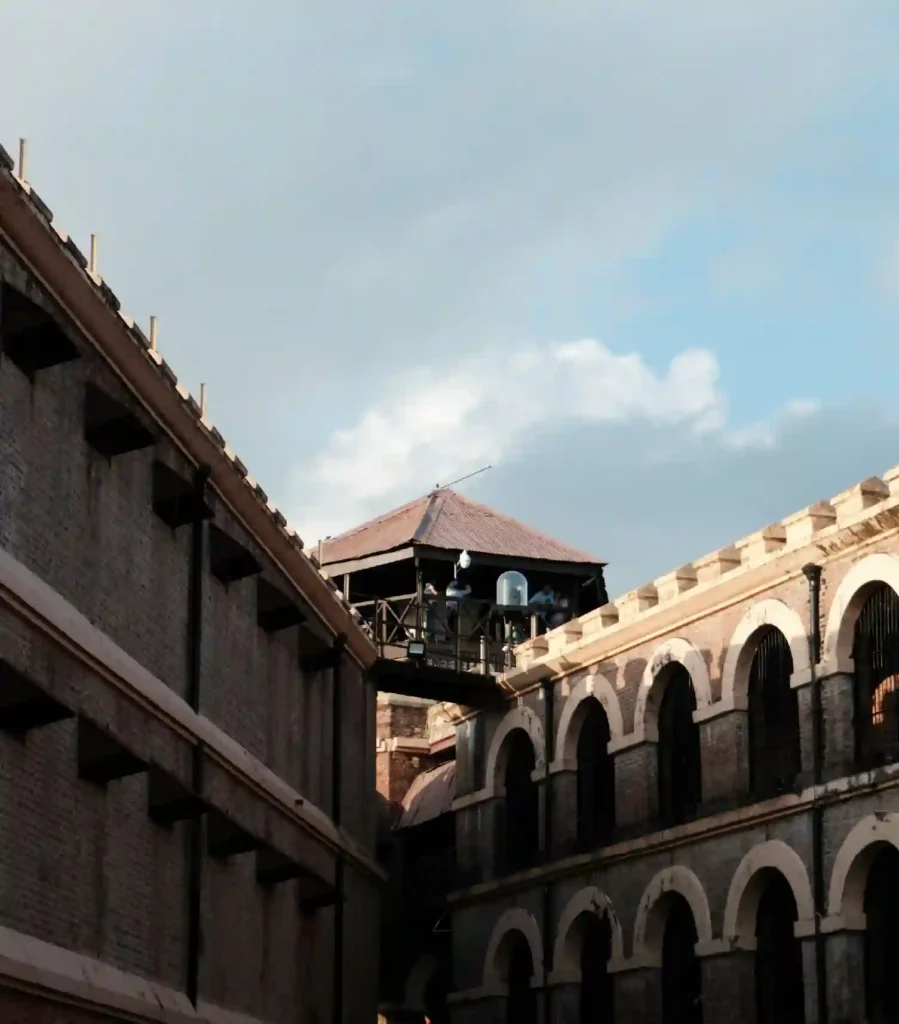 A low-angle shot of the Cellular Jail (Kala Pani) in the Andaman Islands, highlighting its distinct colonial architecture. The image shows the multi-tiered brick corridors with large arches on the right, a plain prison wing on the left, and the central wooden watchtower bridging them under a bright sky.