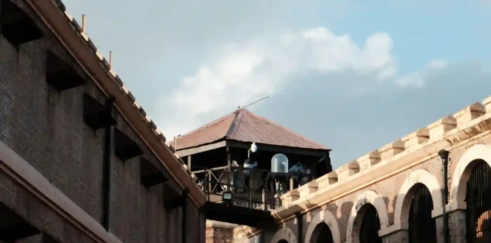 A low-angle shot of the Cellular Jail (Kala Pani) in the Andaman Islands, highlighting its distinct colonial architecture. The image shows the multi-tiered brick corridors with large arches on the right, a plain prison wing on the left, and the central wooden watchtower bridging them under a bright sky.