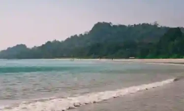 Gentle white waves lapping the shores of Radhanagar Beach on Havelock Island, Andaman, with a wide sandy coastline and lush green tropical forests in the background.