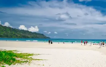 A sunny view of Radhanagar Beach with wide white sand, scattered visitors, turquoise ocean water, and green hills under a bright blue sky.