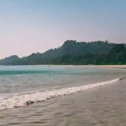 Gentle white waves lapping the shores of Radhanagar Beach on Havelock Island, Andaman, with a wide sandy coastline and lush green tropical forests in the background.