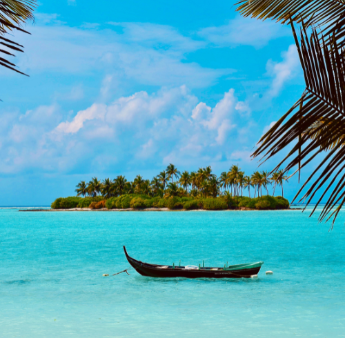 A small wooden boat floating on crystal-clear turquoise water, with a lush tropical island covered in palm trees in the background under a blue sky.