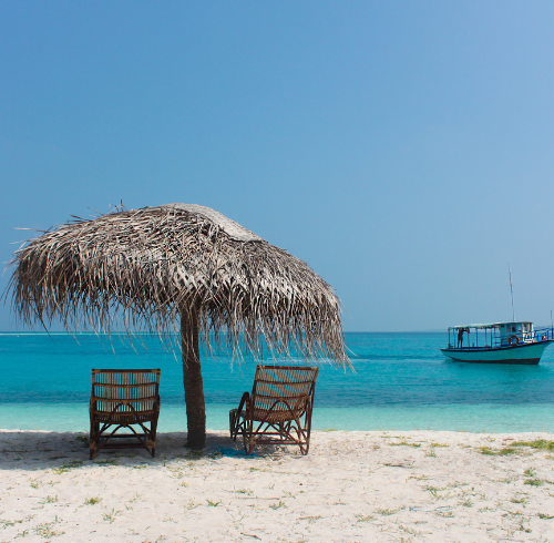 Two wooden lounge chairs positioned under a thatched straw umbrella on a white sand beach, looking out at a turquoise ocean with a boat in the distance.